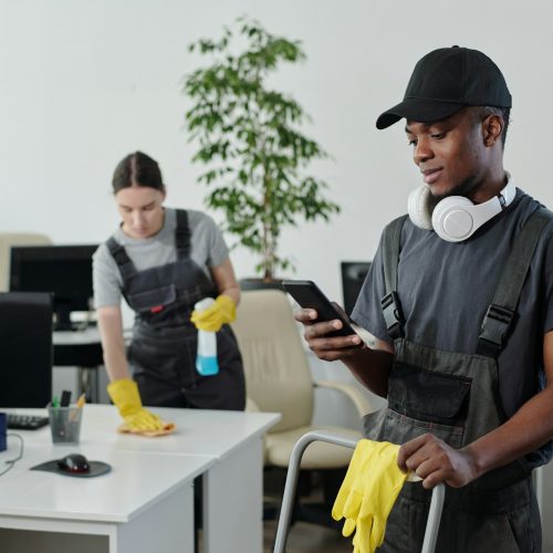 Young male cleaner in workwear choosing music in playlist during work in office